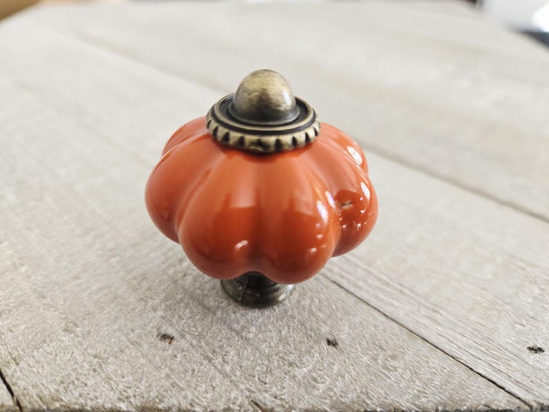 Op de afbeelding: Oranje keramische kastknop in de vorm van een pompoen, met een bronzen basis en bovenkant. De knop heeft een glanzende afwerking en is ontworpen om op een pompoen te lijken.