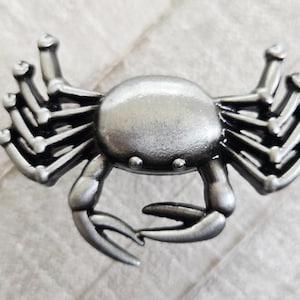 May include: A detailed, silver-toned crab figurine. The crab has a rounded body, multiple legs, and prominent claws. The design features intricate detailing, giving it a realistic appearance. The crab is a decorative item.