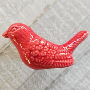 Puede incluir: Pequeña figura de pájaro de cerámica roja con detalles en las alas y la cola. La figura tiene un acabado brillante y está sobre una superficie clara. Es un artículo decorativo.