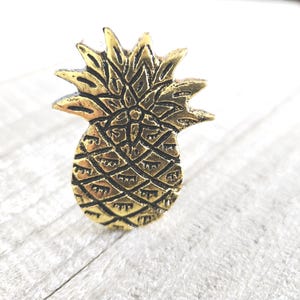 May include: A gold-colored pineapple-shaped decorative object. The pineapple has a textured surface with detailed lines and a crown of spiky leaves. The object is likely a decorative pin or charm.