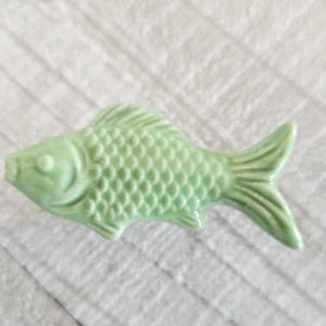 May include: A light green ceramic fish figurine with detailed scales and fins. The fish is a decorative item, possibly for home decor or a collection. The figurine is on a white, textured surface.