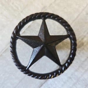 Peut inclure: Élément décoratif en forme d'étoile en fonte noire. L'étoile est centrée dans un cadre circulaire avec une texture de corde. La pièce a un aspect rustique et vieilli, adapté à la décoration western ou de ferme.