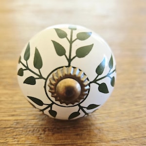 May include: A white ceramic cabinet knob with a botanical design. Dark green leaves and stems are arranged around the knob, with a brass-colored center. The knob is approximately 1.5 inches in diameter.