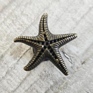 May include: A five-pointed starfish-shaped decorative knob or button featuring a metallic bronze finish with dark recessed grooves that highlight the textured, radial lines extending from the centre to each of the five tapered arms.