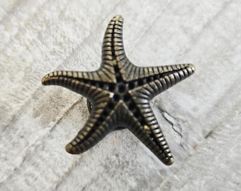 Puede incluir: Un botón decorativo con forma de estrella de mar de cinco puntas, con un acabado en bronce metálico y surcos oscuros que resaltan las líneas radiales texturizadas que se extienden desde el centro hacia cada uno de los cinco brazos.