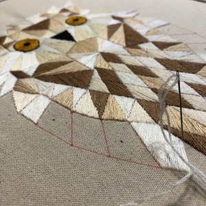 May include: Close-up of a geometric owl embroidery in progress. The owl is made of white, brown, and beige thread on a beige fabric. The needle is threaded with white thread and is being used to stitch the owl's beak.