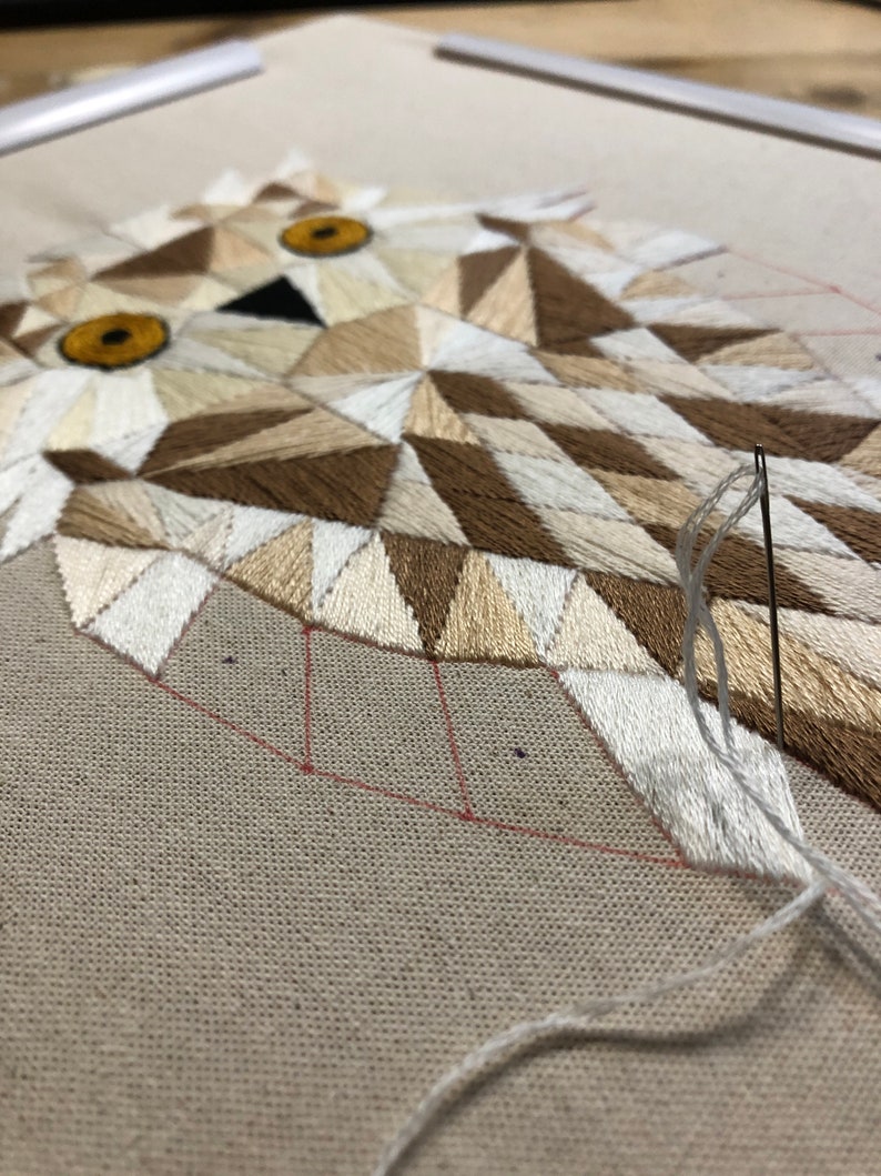 May include: Close-up of a geometric owl embroidery in progress. The owl is made of white, brown, and beige thread on a beige fabric. The needle is threaded with white thread and is being used to stitch the owl's beak.