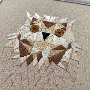 May include: Geometric owl embroidered on a beige fabric with white, brown, and tan thread. The owl's eyes are orange. The background is a red geometric pattern.