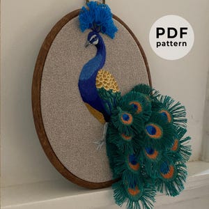 Op de afbeelding: Geborduurde pauw-ontwerp in een houten borduurring. De pauw is blauw, groen en geel met een gedetailleerde staart van veren. Het ontwerp is op een beige stof. De tekst "PDF pattern" staat in de rechterbovenhoek van de afbeelding.