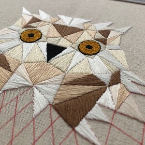 May include: A geometric owl embroidered on a beige fabric with white and brown thread. The owl's eyes are yellow and the beak is black. The fabric has a red grid pattern in the background.