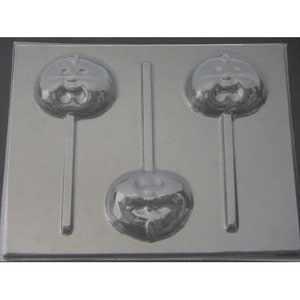 May include: A clear plastic mold for making lollipop candy. The mold has three cavities, each shaped like a pumpkin with a face. The mold is designed to create lollipops with a stick.