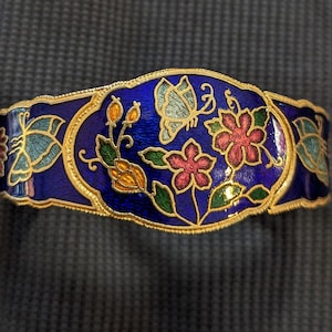 May include: A gold-toned bracelet with a blue enamel background and a floral design featuring butterflies, flowers, and leaves. The flowers are pink, red, and yellow.