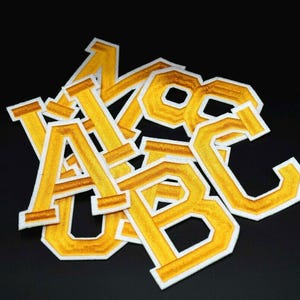 May include: A set of yellow felt letters with white outlines. The letters are A, B, C, K, and S. The letters are arranged in a pile.