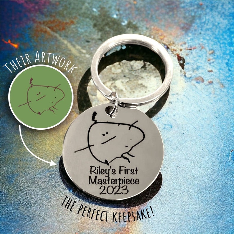 A silver personalised keyring with a child's drawing engraved in black with the wording Riley's First Masterpiece 2023. A small swatch shows the transition from the drawn picture to how it is engraved. Mother's Day gift.