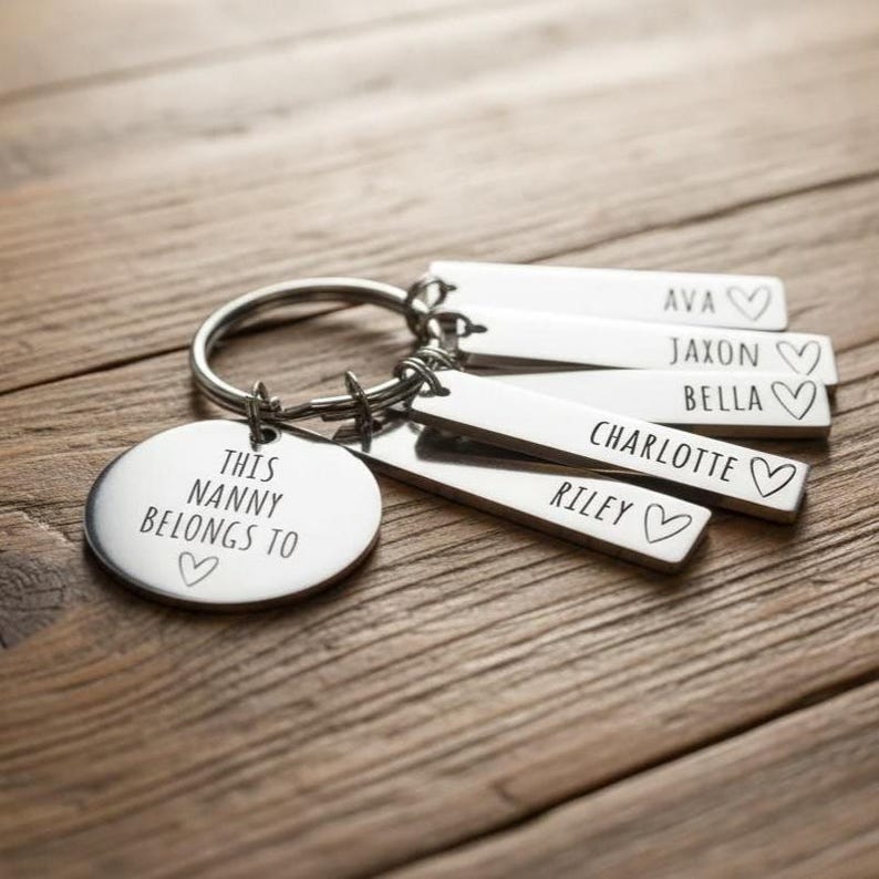 Puede incluir: Un llavero plateado con un dije redondo que dice "THIS NANNY BELONGS TO" y un coraz&oacute;n. Tiene etiquetas rectangulares grabadas con los nombres: AVA, JAXON, BELLA, CHARLOTTE y RILEY, cada una con un coraz&oacute;n. El llavero est&aacute; sobre una superficie de madera.