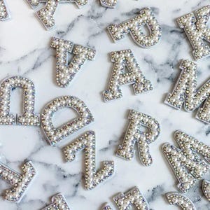 May include: A set of pearl-embellished letters and numbers, including A, B, C, D, E, F, G, H, I, J, K, L, M, N, O, P, Q, R, S, T, U, V, W, X, Y, Z, 1, 2, 3, 4, 5, 6, 7, 8, 9, and 0. The letters and numbers are white and have a pearl-like finish.