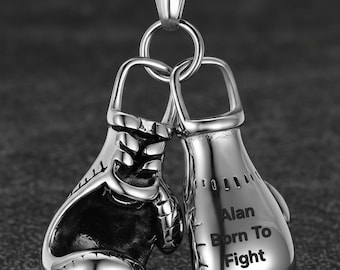 Könnte beinhalten: Ein silberfarbener Metallanhänger mit zwei Miniatur-Boxhandschuhen, die an einer passenden Kette hängen. Ein Handschuh ist mit dem Text 'Alan Born To Fight' in einer klaren, dunklen Schrift graviert. Das Design zeigt detaillierte Nähte.