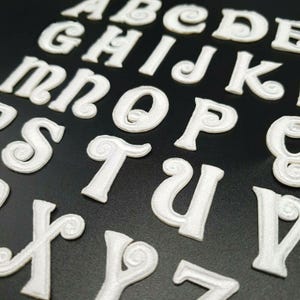 May include: White felt letters of the alphabet with a decorative swirl design. The letters are arranged in a random order on a black background.