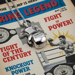 Puede incluir: Un colgante de guante de boxeo plateado con el texto grabado "Andrew Born To Fight" cuelga de una cadena plateada. El colgante descansa sobre un p&oacute;ster de boxeo vintage con las palabras "Ring Legend" y "Fight of Power!"