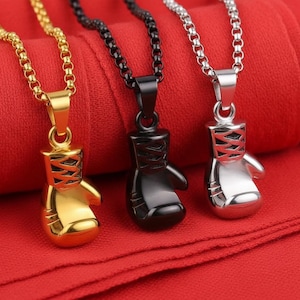 Puede incluir: Tres collares con colgantes de guantes de boxeo. Uno es dorado, otro negro y otro plateado. Cada colgante tiene un dise&ntilde;o detallado y cuelga de una cadena a juego. El fondo es de tela roja.