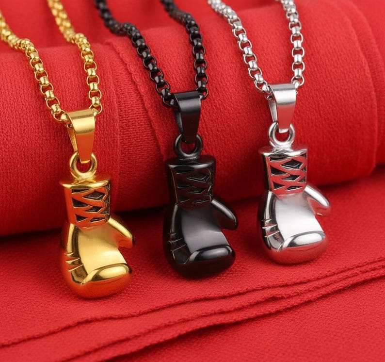 Puede incluir: Tres collares con colgantes de guantes de boxeo. Uno es dorado, otro negro y otro plateado. Cada colgante tiene un dise&ntilde;o detallado y cuelga de una cadena a juego. El fondo es de tela roja.