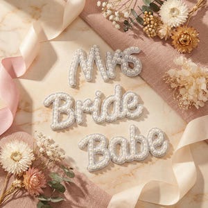 May include: Three decorative, cursive word cutouts: "Mrs", "Bride", and "Babe". Each word is made of white pearls with a silver border. The words are arranged on a pink cloth with dried flowers and ribbons.