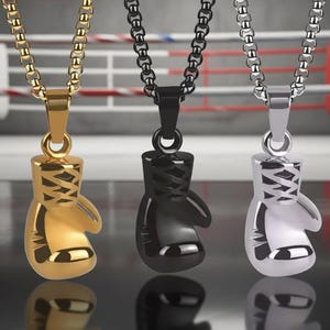 Puede incluir: Tres collares con colgantes de guantes de boxeo en oro, negro y plata. Cada colgante est&aacute; detallado con cordones y cuelga de una cadena a juego. El fondo muestra un ring de boxeo.