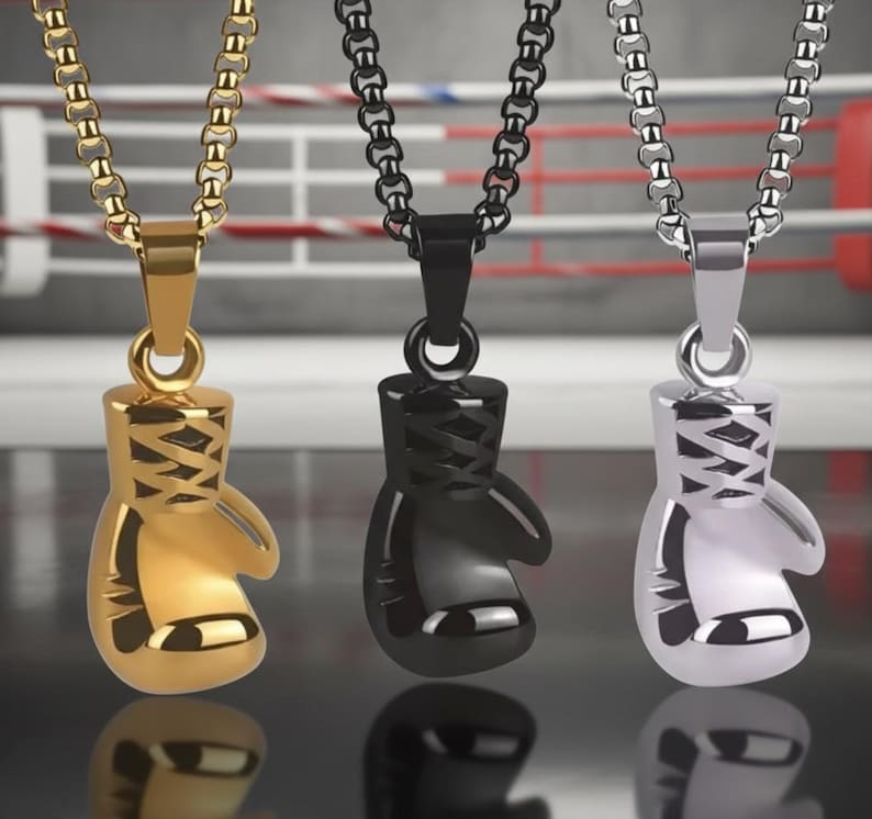 Puede incluir: Tres collares con colgantes de guantes de boxeo en oro, negro y plata. Cada colgante est&aacute; detallado con cordones y cuelga de una cadena a juego. El fondo muestra un ring de boxeo.