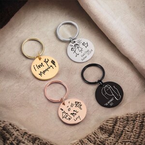 May include: Four silver keychains with round metal tags. Each tag features a different child's drawing and inscription. One tag says "i love You Mummy", another says "Park Day Joshua - 2024", another says "Lucy", and the last one says "THE MERMAID 2021".