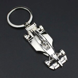 May include: Silver-toned metal keychain featuring a detailed model of a race car. The car has a driver figure inside and is attached to a silver key ring.