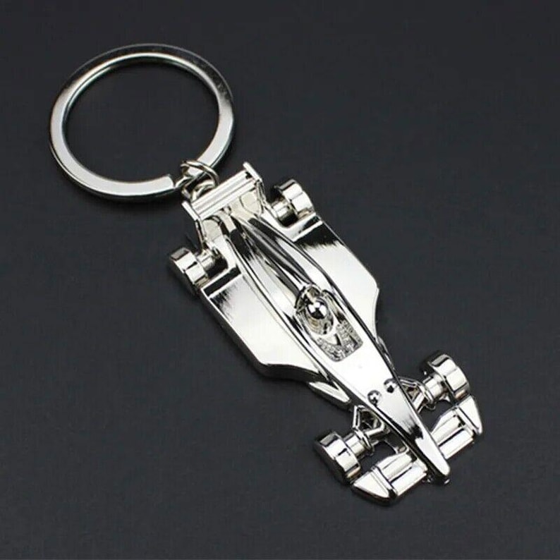 May include: Silver-toned metal keychain featuring a detailed model of a race car. The car has a driver figure inside and is attached to a silver key ring.
