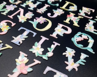 Könnte beinhalten: Ein Set gestickter Bügelbilder mit den Buchstaben des Alphabets von A bis Z. Jeder Buchstabe ist mit pastellfarbenen Blumen und Blättern verziert.