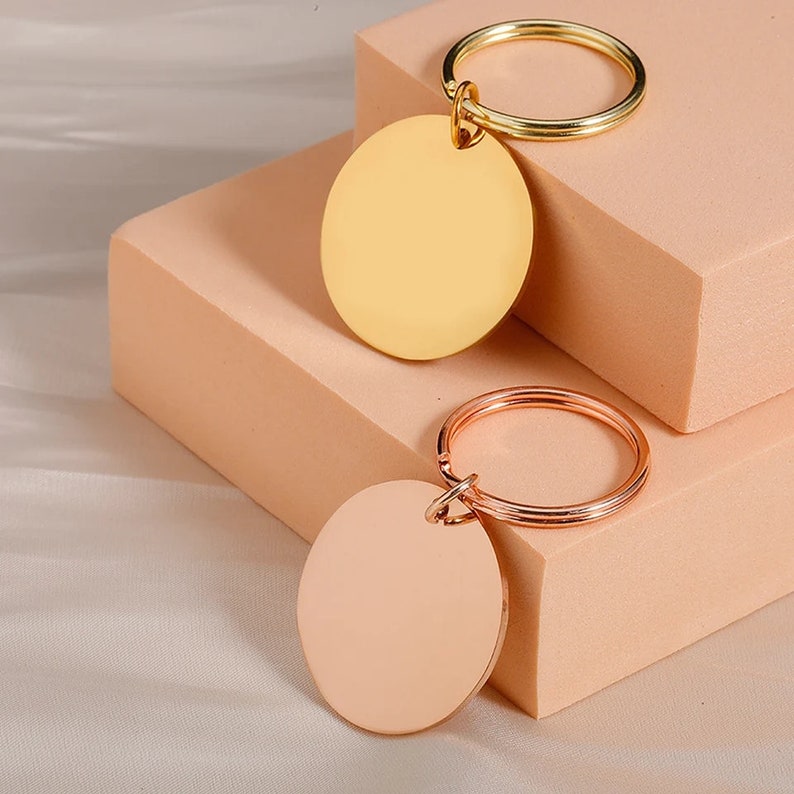 May include: Two blank round metal keychains, one gold and one rose gold, with a split ring attached to each. The keychains are on a light pink surface.