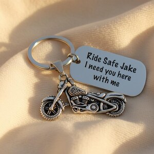 May include: Silver motorcycle keychain with a matching tag. The tag reads "Ride Safe Jake I need you here with me". The keychain is on a soft, beige fabric.