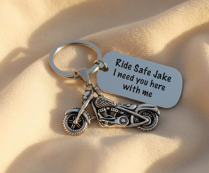 May include: Silver motorcycle keychain with a matching tag. The tag reads "Ride Safe Jake I need you here with me". The keychain is on a soft, beige fabric.