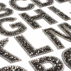 May include: A set of silver glitter letters of the alphabet, A through J and L through O, on a white background.