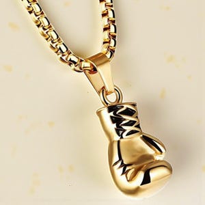 Puede incluir: Collar con colgante dorado con un dije de guante de boxeo. El dije tiene detalles negros y est&aacute; suspendido de una cadena dorada. El collar se muestra sobre un fondo claro.