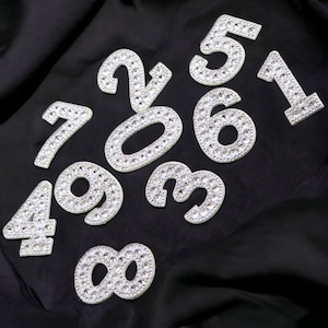 May include: A selection of rhinestone-covered numbers, including 1, 2, 3, 4, 5, 6, 7, 8, and 9, are shown against a black fabric backdrop. The numbers are white with a sparkling, gem-like appearance.