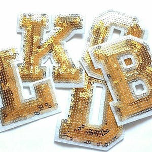 Può includere: Quattro lettere dorate con paillettes, L, K, B e D, con supporto in feltro bianco. Le lettere sono disposte in modo informale, leggermente inclinate.