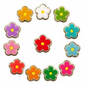 May include: A collection of colorful, embroidered flower patches. Each flower has a textured, plush center in shades of orange, white, green, purple, pink, red, and blue, with a gold-colored trim. The patches are arranged in a circular pattern.