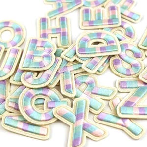 Op de afbeelding: Een verzameling kleurrijke, geborduurde alfabetletters met een pastel gestreept patroon. De letters zijn gemaakt van stof en hebben een witte achtergrond.