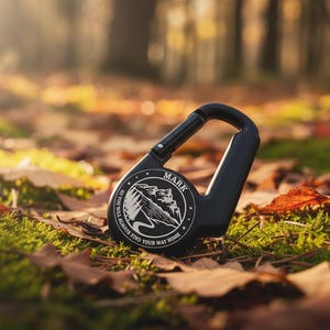 Op de afbeelding: Een zwarte karabijnhaak met een rond embleem met een illustratie van een berglandschap en de tekst "MARK" en "SO YOU WILL ALWAYS FIND YOUR WAY HOME" in het wit gedrukt. De karabijnhaak heeft een veerbelaste sluiting en een gladde, matte afwerking.