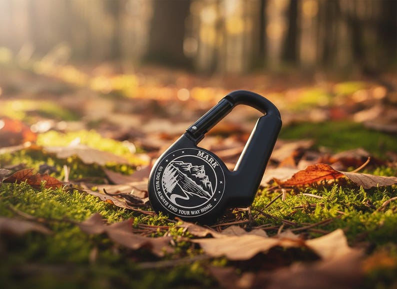 Op de afbeelding: Een zwarte karabijnhaak met een rond embleem met een illustratie van een berglandschap en de tekst "MARK" en "SO YOU WILL ALWAYS FIND YOUR WAY HOME" in het wit gedrukt. De karabijnhaak heeft een veerbelaste sluiting en een gladde, matte afwerking.