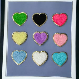 Op de afbeelding: Negen hartvormige patches met een gouden rand. De patches zijn in verschillende kleuren, waaronder groen, zwart, roze, geel, paars, lichtroze, lichtblauw, wit en turquoise.