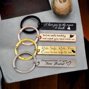 May include: Four silver keychains with different engraved messages. The messages are "I love you to the moon & back", "Drive safe Daddy We need you here with us", "Ride Safe, Ride Free Come home safe to me", and "Congratulations on your New Home!"