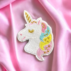 May include: Embroidered unicorn patch with a white head, gold horn, and colourful mane in pink, blue, and yellow. The unicorn has closed eyes and a smiling mouth. The patch is on a pink satin background.