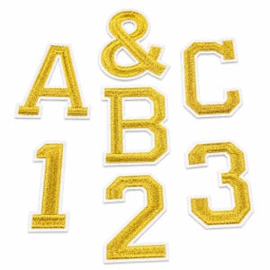May include: A collection of gold embroidered patches featuring the letters A, B, and C, the numbers 1, 2, and 3, and an ampersand symbol. Each patch has a textured metallic gold finish with a clean white border, arranged in a scattered layout.