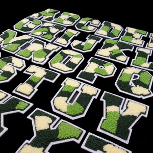 May include: A set of 26 iron-on patches featuring the letters of the alphabet in a green and white camouflage pattern. The patches are outlined in white and have a fuzzy texture.