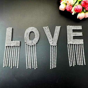 May include: Four rhinestone letters spelling out the word "LOVE" with dangling rhinestone fringe. Each letter is silver and has a sparkly finish.