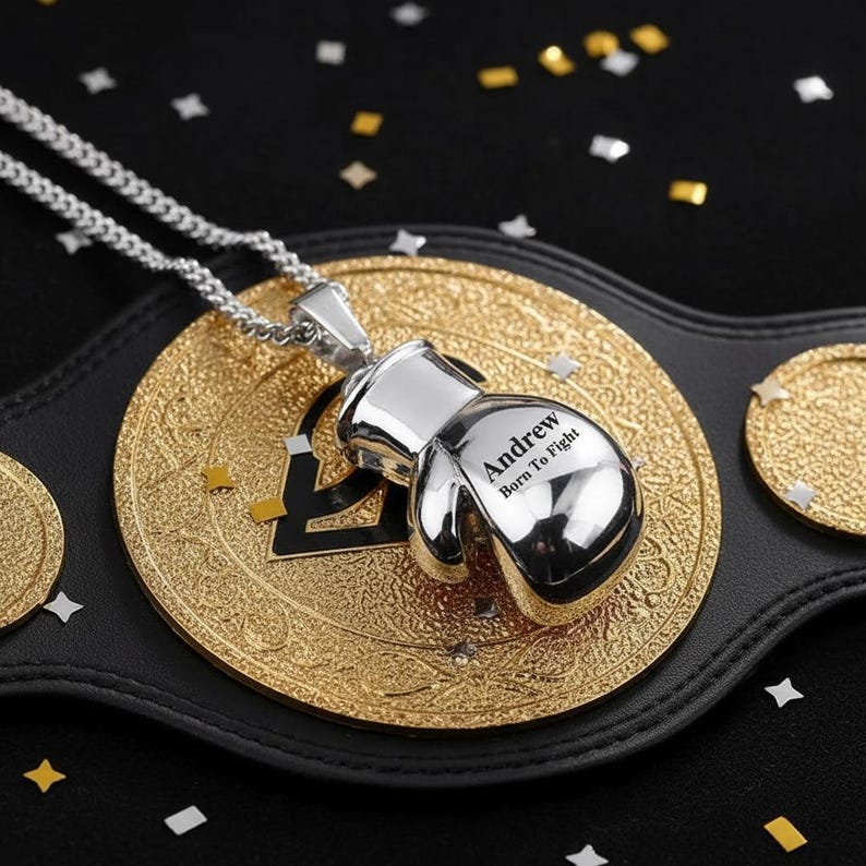 Puede incluir: Un collar con colgante de guante de boxeo plateado con el texto "Andrew Born To Fight" grabado. El colgante cuelga de una cadena plateada, sobre un cintur&oacute;n de campeonato dorado.